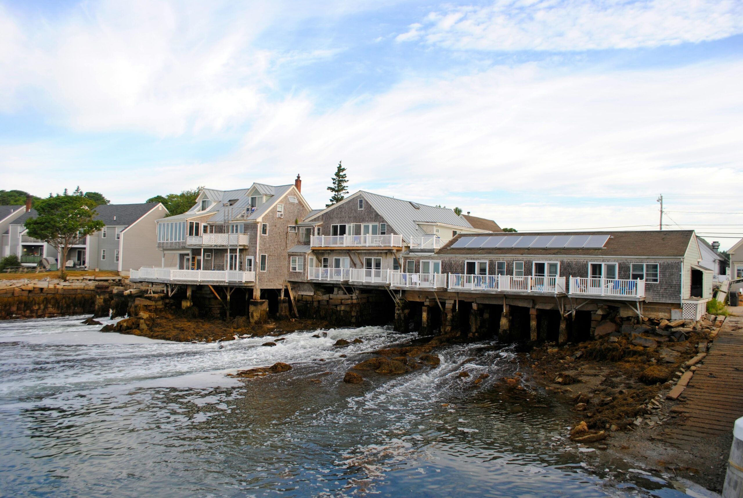 Exteriors of aged residential houses located on rocky coast of wavy ocean against cloudy blue sky